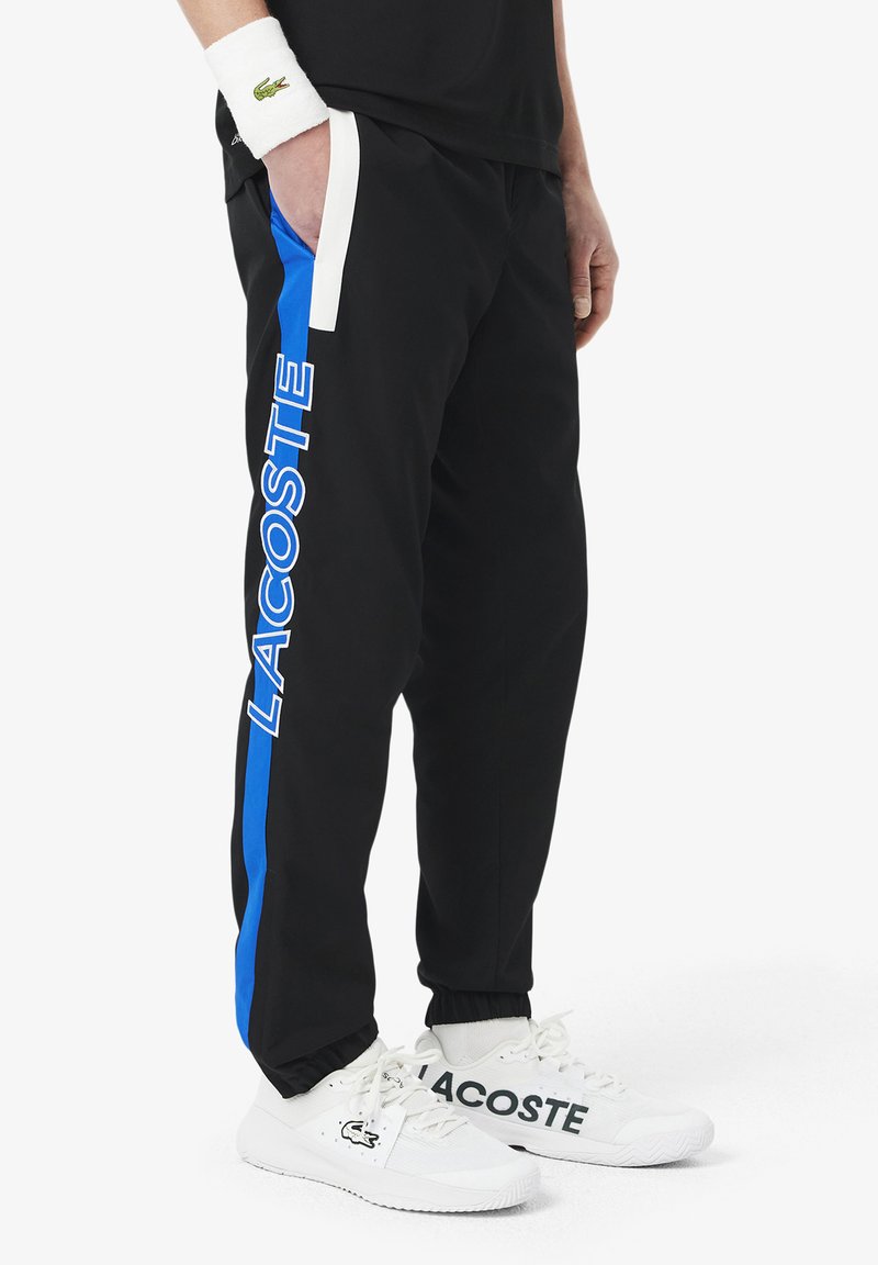 Zwarte joggingbroeken met blauwe zijkanten en "LACOSTE" merknaam. Gemaakt van zacht materiaal met een elastische tailleband en boorden bij de enkels.