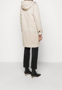 Licht beige jas met een capuchon, met een eenvoudig ontwerp en knoopdetails. Gedragen over zwarte nauwsluitende broek en zwarte enkellaarsjes.