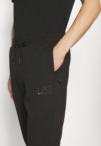 Black sweatpants in soft fabric featuring a drawstring waistband and side pockets. Logo printed on the front leg. Simple and functional design.