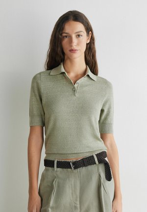 Frau mit langen braunen Haaren trägt ein hellgrünes, kurzärmeliges Strickhemd mit Kragen, hellgrüne Hose mit hoher Taille und schwarzen gewebten Gürtel.