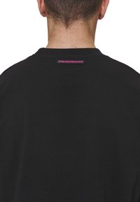 Black t-shirt made of soft cotton, featuring a crew neck and a logo tag at the back neck in pink and black colors. Simple, relaxed fit.