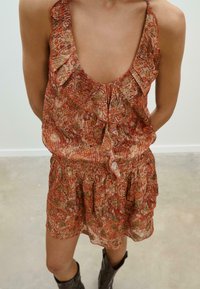 Abito rosso e oro con motivo paisley, dotato di spalline arricciate e vita arricciata, realizzato in tessuto leggero. Stivali marroni completano il look.