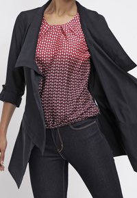 Giacca nera aperta davanti sopra una blusa rossa a fantasia con pois bianchi. La blusa ha un colletto arricciato. Indossata con jeans in denim scuri.