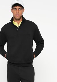 Μαύρο φούτερ με φερμουάρ και όρθιο γιακά, με λογότυπο στο στήθος και ριγέ μανσέτες, σε συνδυασμό με μαύρο παντελόνι και λευκό καπέλο.