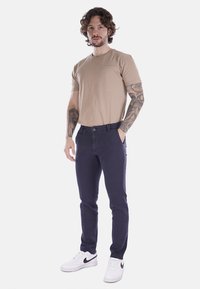 T-shirt in cotone beige con taschino, abbinato a pantaloni slim-fit blu navy e sneakers bianche con dettagli neri.