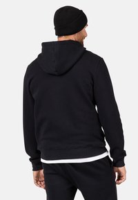 Schwarzer Kapuzenpullover mit Kordelzug, gerippten Bündchen und Saum. Weicher, strukturierter Stoff; getragen mit einer schwarzen Mütze. Schlichtes Design, keine sichtbaren Muster.