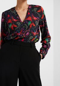 Blouse à manches longues à motif floral avec un col en V profond, présentant un fond noir et des motifs rouges, verts et violets éclatants. Associée à un pantalon noir taille haute.