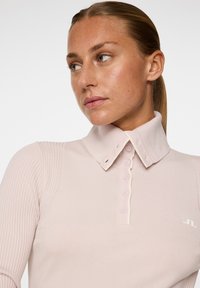 Jasnoróżowa, długorękawowa koszulka polo o prążkowanej teksturze, z zagiętym kołnierzykiem, listwą z guzikami i małym białym logo na piersi.