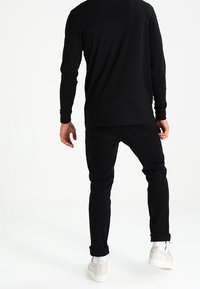 Mies seisoo selkä kameraan, päällään musta pitkähiha-tee paita, mustat farkut, valkoiset sukat ja vaaleat lenkkitossut valkoista taustaa vasten.