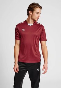 Dunkelroter Sporttrikot mit kurzen Ärmeln, V-Ausschnitt, weißen Akzenten auf den Schultern und „hummel“-Logo auf der Brust. Schwarze Hose.