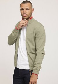 Cardigan zippé en coton vert avec des accents rouges, col et poignets côtelés, et un petit logo. Porté sur un t-shirt blanc et un jean foncé.