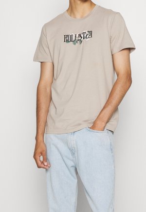 Beige katoenen T-shirt met de tekst "HOLLISTER" en bloemaccenten. Ronde hals, korte mouwen en een losse pasvorm. Gecombineerd met lichtblauwe jeans.