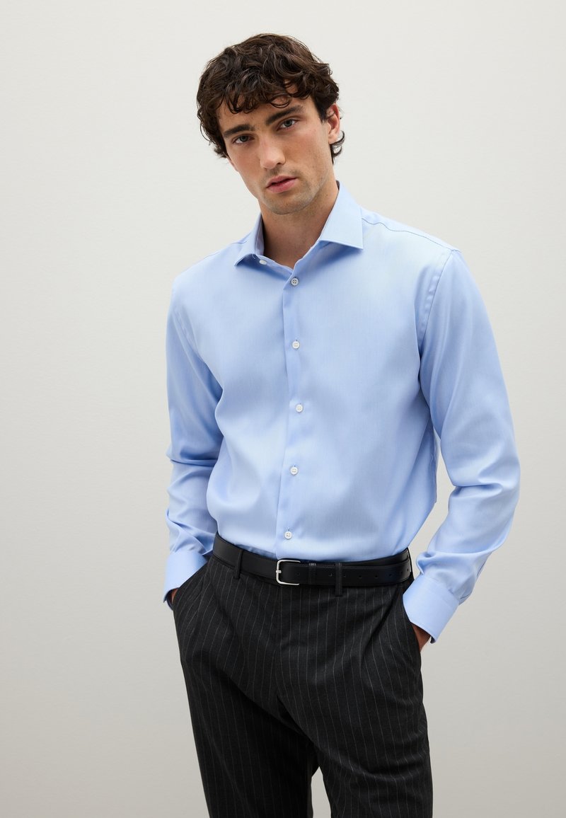 Giovane vestito con una camicia blu chiaro infilata in pantaloni scuri a righe con una cintura nera, in piedi con le mani nelle tasche.