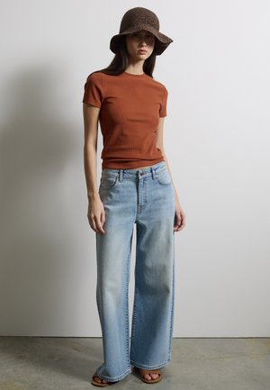 Vrouw die een bruin kortärmelig shirt, lichtblauwe wijde jeans, bruine sandalen en een bruine gebreide bucket hat draagt, staand tegen een effen muur.