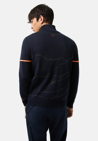 Dunkelblaues Strickpullover mit einem hohen Kragen, der ein dezentes Bergmuster in Hellgrau und einen orangefarbenen Streifen am Ärmel aufweist.