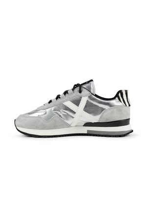 Zapatilla baja en plata y gris con cruz blanca lateral, cordones negros, lengüeta del talón a rayas en negro y blanco, y suela negra y blanca.