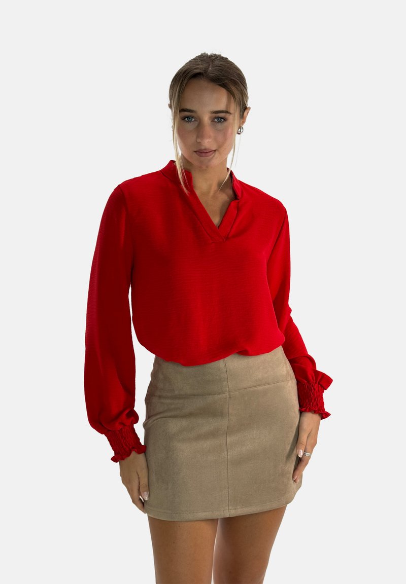 Blusa rossa a maniche lunghe con scollatura a V e polsini arricciati, abbinata a una mini gonna in suede beige. Tessuto liscio con sottili dettagli di design.