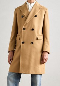 Cappotto doppiopetto in lana color cammello, con bottoni neri, collo a revers, tasche laterali e taglio dritto. Indossato sopra una camicia bianca e jeans blu.