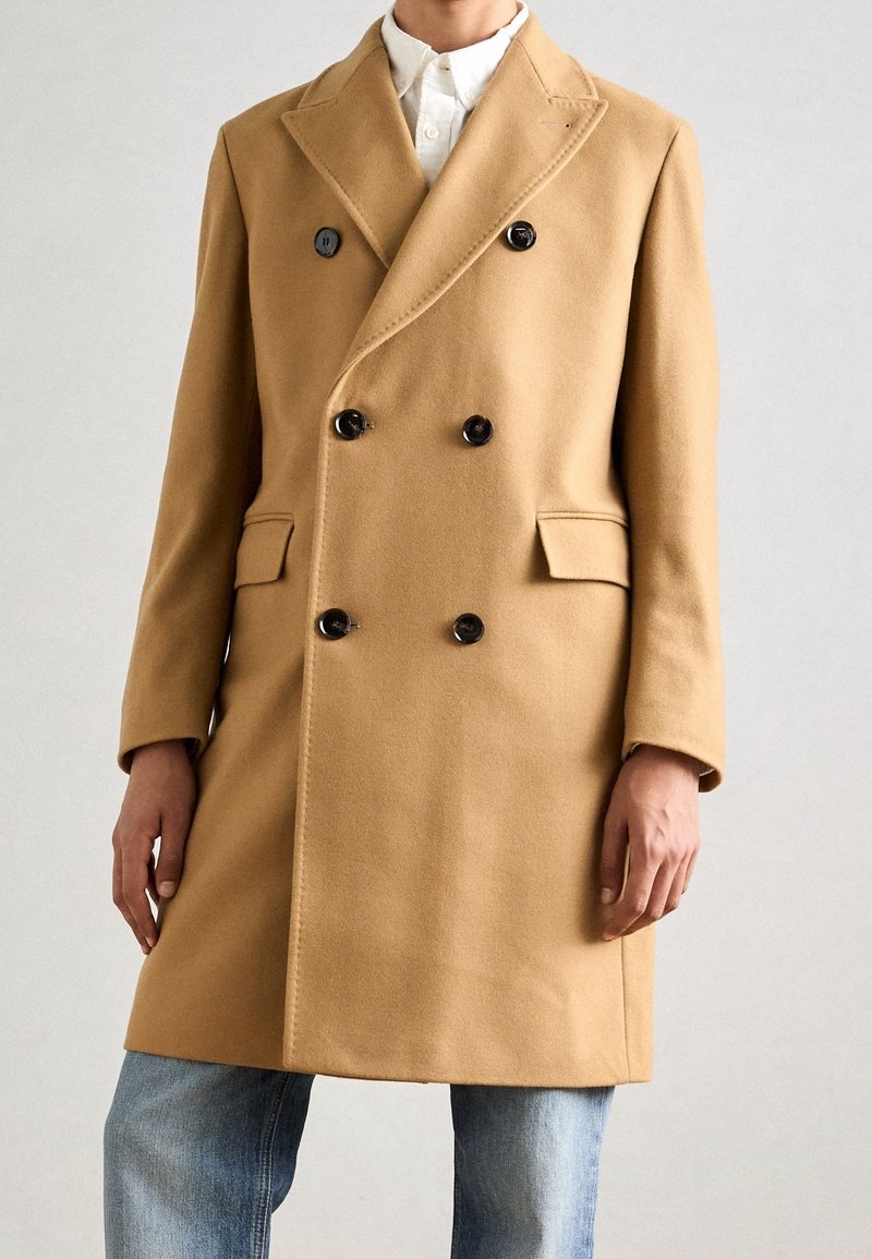 Cappotto doppiopetto in lana color cammello, con bottoni neri, collo a revers, tasche laterali e taglio dritto. Indossato sopra una camicia bianca e jeans blu.