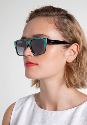 Donna con capelli corti biondi che indossa grandi occhiali da sole verdi e neri con motivo e un top bianco senza maniche testurizzato.