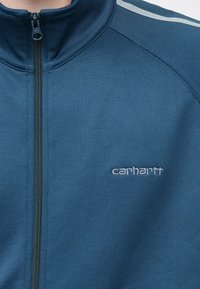 Ζακέτα με φερμουάρ σε σκούρο μπλε χρώμα και ψηλό γιακά, με γκρι ρίγες τόνου και μικρό κεντητό λογότυπο "Carhartt" στο κάτω δεξί μέρος.