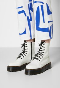 Botas de cuero blanco con cordones y suela gruesa en negro y naranja, que cuentan con una cremallera lateral y un detalle de costura pronunciada.