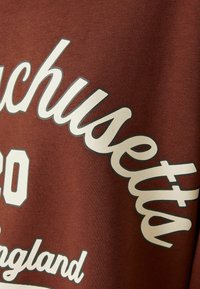 Sweatshirt marron arborant un grand texte blanc indiquant "Massachusetts 20" et "Angleterre." Le texte présente un léger effet d'ombre pour plus de profondeur.