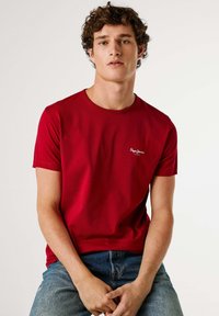 Rood T-shirt met korte mouwen, gemaakt van katoen, met een ronde hals en een klein wit merklogo op de linkerborst. Draagt bij een blauwe spijkerbroek.