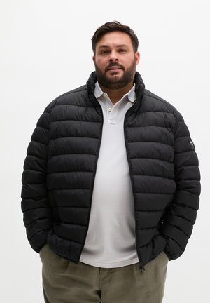 Mann mit Bart, der eine schwarze Daunenjacke über einem weißen Poloshirt und khakifarbenen Hosen trägt, steht mit den Händen in den Taschen vor einem einfarbigen Hintergrund.