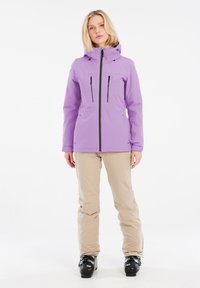 Lila wasserdichte Jacke mit Kapuze, ausgestattet mit Reißverschlusstaschen und Belüftungsdetails, kombiniert mit beigen Hosen und schwarzen Skischuhen.
