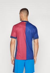 Maglia da calcio con sezioni verticali rosse e blu, maniche corte e scollo rotondo. Presenta un dettaglio a striscia orizzontale sulla parte posteriore del collo.