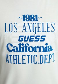 T-shirt bianco con testo blu: "1981 LOS ANGELES GUESS California. ATHLETIC DEPT." Testo in vari stili e dimensioni di carattere.