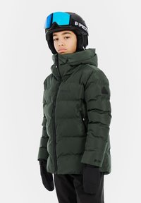Grøn pufferjakke med høj krave og lynlås, med et sort logo-patch på ærmet. Bæres med sorte handsker og snebriller.