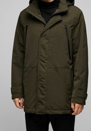 Homme portant un manteau d'hiver à capuche de couleur vert olive avec deux poches poitrine zippées et rabat sur la fermeture éclair frontale.
