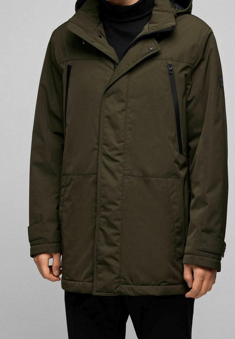 Homme portant un manteau d'hiver à capuche de couleur vert olive avec deux poches poitrine zippées et rabat sur la fermeture éclair frontale.