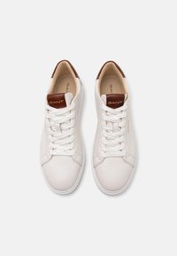 Baskets en cuir blanc avec bout rond, lacets blancs, et accent en cuir marron au niveau du talon. Logo de la marque en or sur la languette. Texture lisse.