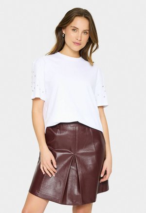 T-shirt blanc à manches courtes avec des embellissements décoratifs sur les manches, associé à une jupe plissée en simili cuir bordeaux.