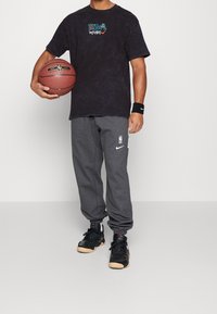 T-shirt noir tie-dye avec le logo de la NBA, pantalon de survêtement gris avec logo, baskets noires, tenant un ballon de basket, bracelet au poignet gauche.