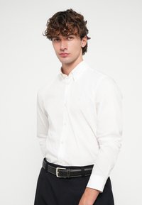 Λευκό πουκάμισο με μακριά μανίκια, γιακά με κουμπιά και διακριτικό λογότυπο στο στήθος, σε συνδυασμό με μαύρη ζώνη και σκούρο παντελόνι.