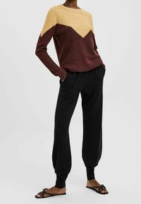 Maglione in oro e bordeaux con un motivo a zigzag; maniche lunghe e scollo rotondo. Pantaloni neri a vestibilità ampia e sandali flat.