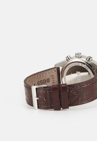 Bracelet de montre en cuir marron avec texture crocodile, boucle en argent et boîtier en acier inoxydable. Résistant à l'eau jusqu'à 5 bars, nom de la marque visible au dos.