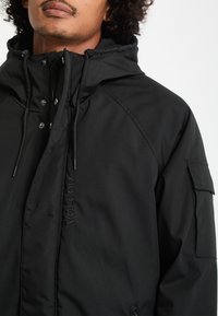 Homme portant une veste à capuche noire avec des cordons et une poche zippée sur le haut de la manche, regardant devant avec une expression neutre.