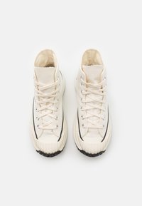 Wysokie sneakersy z kremową cholewką z płótna, czarną gumową podeszwą i białymi sznurowadłami, z zaokrąglonym noskiem i widocznymi detalami przeszyć.