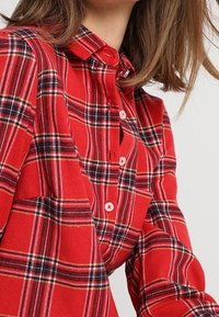 Chemise à carreaux rouges en coton. Elle présente un design à boutons, un col et trois boutons. Des rayures noires et jaunes sont présentes dans le motif.