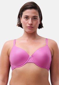 Frau mit kurzem dunklem Haar, die einen glatten, pinkfarbenen Bügel-BH mit verstellbaren Trägern vor einem schlichten weißen Hintergrund trägt.