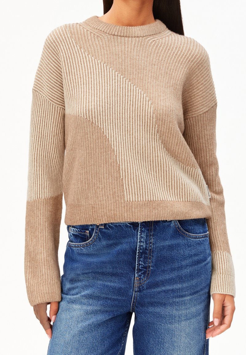 Femme portant un pull en maille côtelée beige et crème avec des motifs abstraits, associé à un jean bleu, montrée du cou aux hanches.