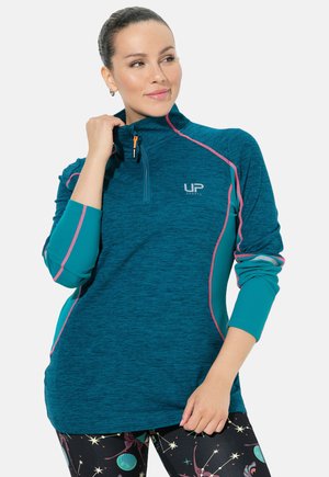 Türkiser Sportpullover mit Reißverschlusskragen, kontrastierenden pinken Nähten und Logo. Strukturierter Stoff und lange Ärmel. Darunter eine Reihe gemusterter Leggings.