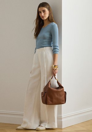 Ženska v svetlo modrem puloverju in širokih belih hlačah, ki drži rjavo usnjeno torbico, stoji v notranjosti ob bež in beli steni.