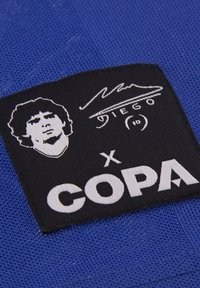 Tela azul con una etiqueta negra que presenta un gráfico blanco de un rostro y el texto "Diego" y "COPA", indicando un producto relacionado con el fútbol.