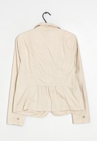 Veste légère beige avec une taille peplum, manches longues et poignets boutonnés. Dotée d'un col classique et d'un tissu texturé. Suspendue à un crochet.